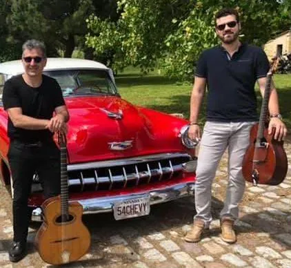 Deux musiciens du groupe avec leurs guitares devant une voiture ancienne rouge