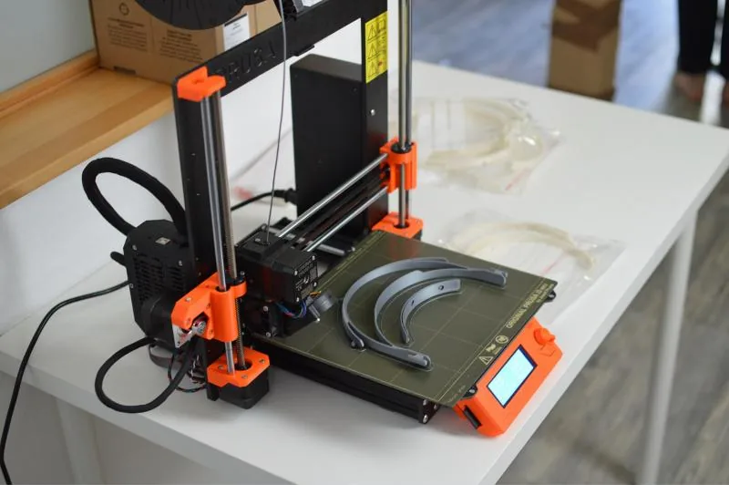 Imprimante Original Prusa i3 MK3, cadre noir et pièces orange, avec des montures de visières sur son plateau